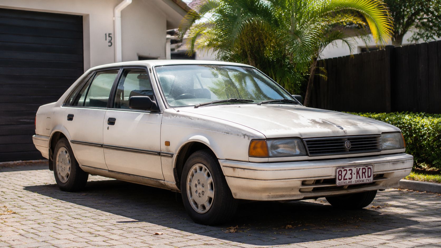 An old white sedan car parked on a paved driveway in front of a residential garage.